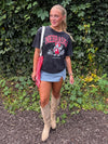 A woman stands outdoors in front of a leafy green hedge and mulch, wearing a faded black Nebraska graphic t-shirt, a denim skirt, beige over-the-knee boots, and carrying a red-and-white checkered shoulder bag.