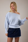 Woman wearing a light blue off-shoulder sweater and dark denim skirt against a plain background