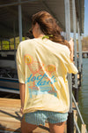 Person wearing a yellow t-shirt with a graphic design, standing on a dock by water.