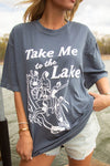 Person wearing a blue t-shirt with 'Take Me to the Lake' text and graphics by a lake.