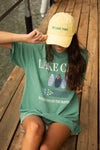Person wearing a green 'Lake Life' t-shirt and yellow cap on a wooden dock by a lake.