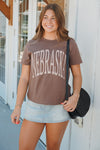 A woman wearing a brown t-shirt with 'NEBRASKA' printed in white arched letters, paired with a light denim mini skirt and a black woven shoulder bag, standing outdoors against a white building wall.