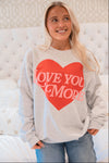 Woman wearing a gray sweatshirt with a red heart and 'LOVE YOU MORE' text, sitting on a white couch.