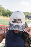 A person outdoors at a sports field wearing a white baseball cap with 'CHOOSIN' NEBRASKA' embroidered in brown on the front and a camouflage-patterned brim.