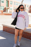 Person wearing a light gray sweatshirt with 'MAVERICKS' text, black shorts, and white sneakers, standing outdoors.