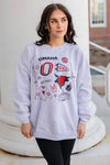 Woman wearing a light blue sweatshirt with 'Omaha' and sports team graphics, standing outdoors.