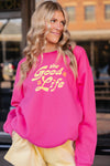 Person wearing a bright pink sweatshirt with 'the Good Life' text, standing outdoors.