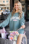 Woman in a teal sweatshirt with 'Nebraska' printed on it, standing next to a bicycle with flowers in an urban setting.